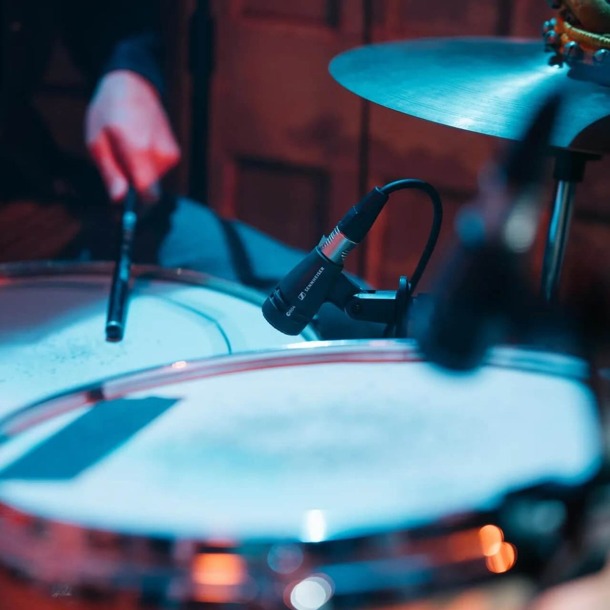 A close-up shot of a Sennheiser e 604 Dynamic Cardioid Instrument Microphone mounted on a stand in front of a drum kit, with a drummer's hand holding a drumstick blurred in the background under blue stage lighting.