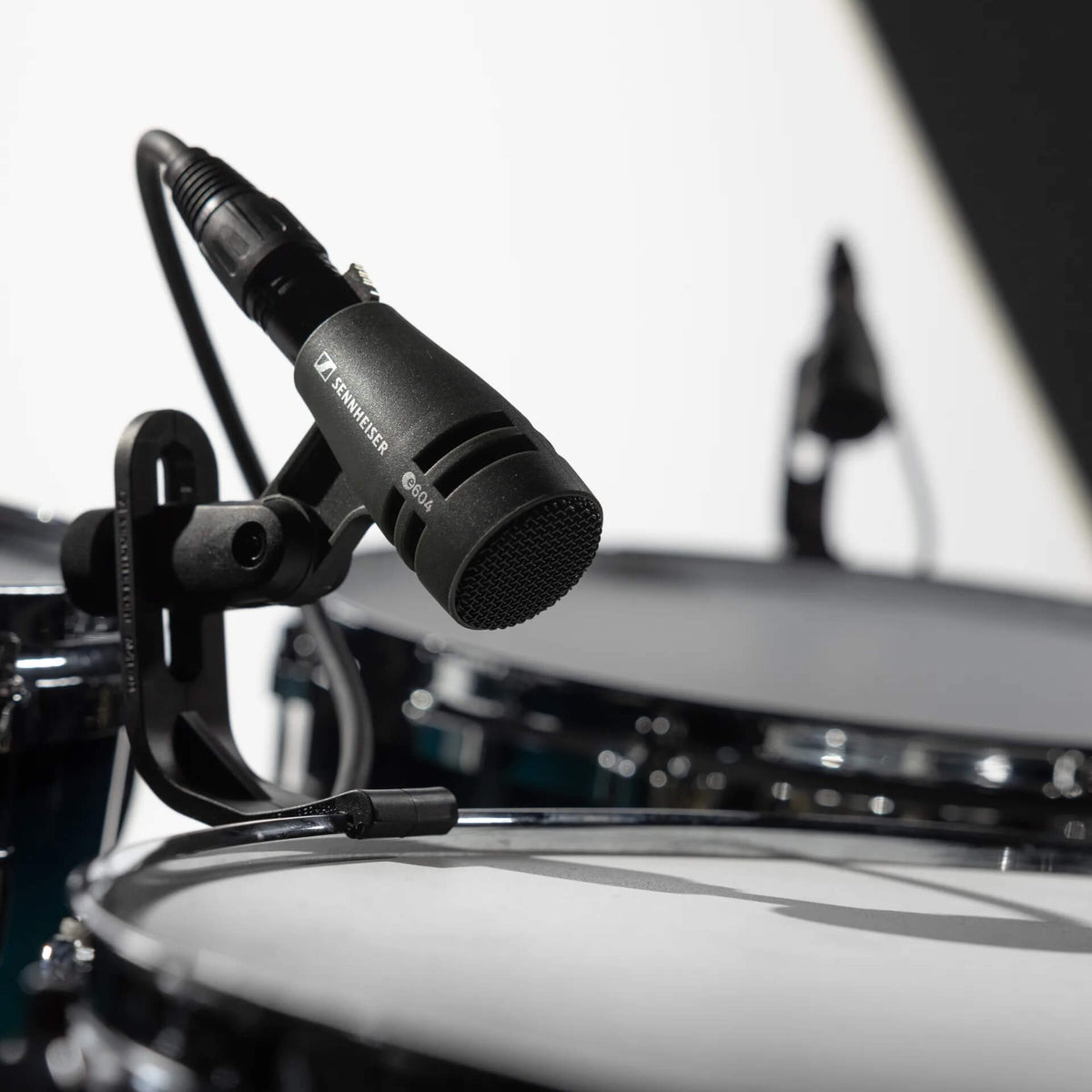 A close-up of a Sennheiser e 604 Dynamic Cardioid Instrument Microphone mounted on a black stand, positioned over a white drum head with other drums blurred in the background.