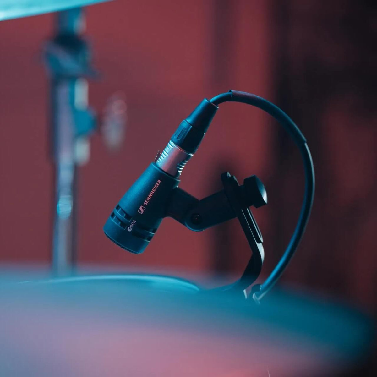 A Sennheiser e 604 Dynamic Cardioid Instrument Microphone mounted on a black clip with an attached cable, shown in a studio setting with warm red and cool blue lighting.